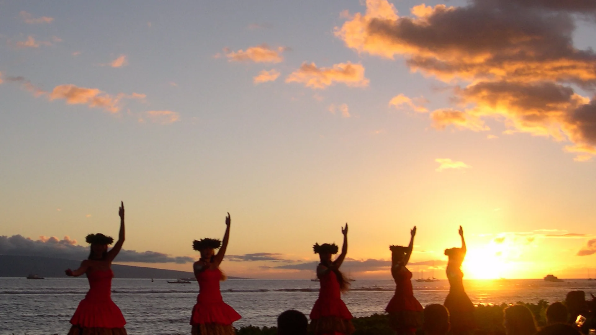 Tancerki hula w czerwonych sukienkach wznoszą ramiona w sylwetce na tle płonącego pomarańczowego zachodu słońca nad oceanem, a goście luau siedzą przy stolikach na pierwszym planie