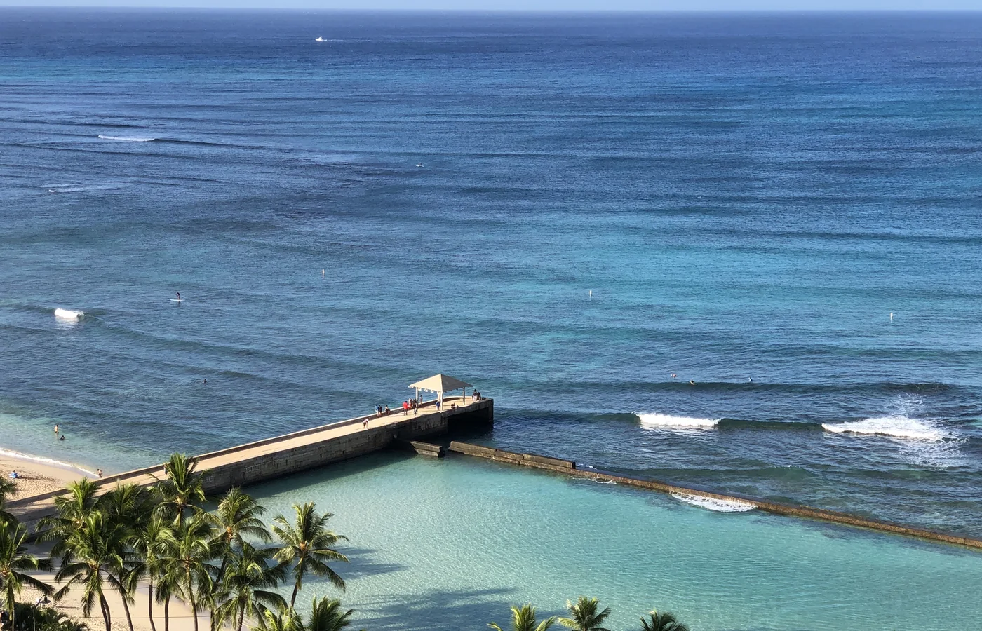 Turkusowa laguna i otwarty Pacyfik widziane z wysokiego balkonu nad Waikiki, z kamiennym pomostem, surferami i palmami na pierwszym planie
